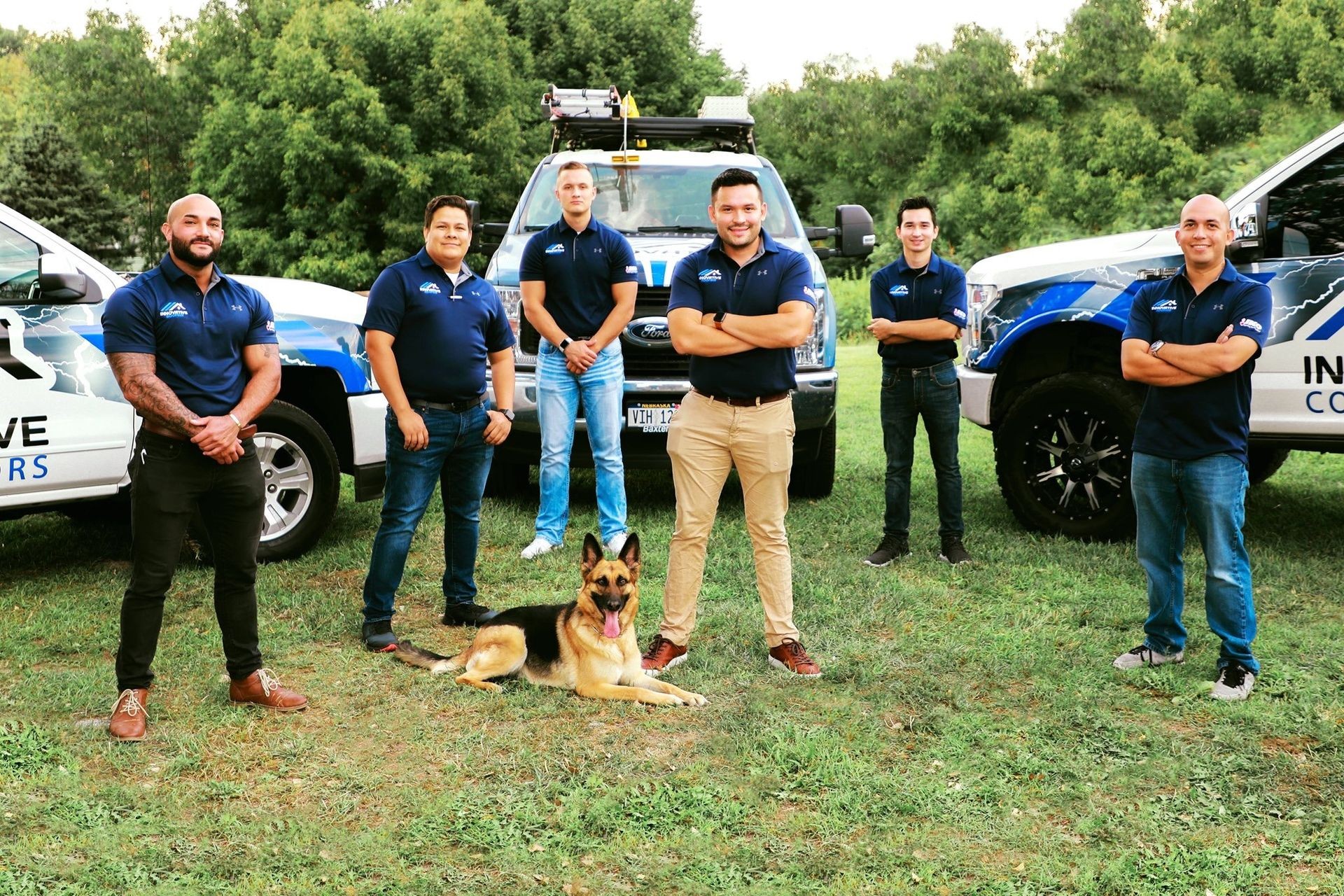 Group of men in matching shirts standing with a dog in front of branded company trucks outdoors.